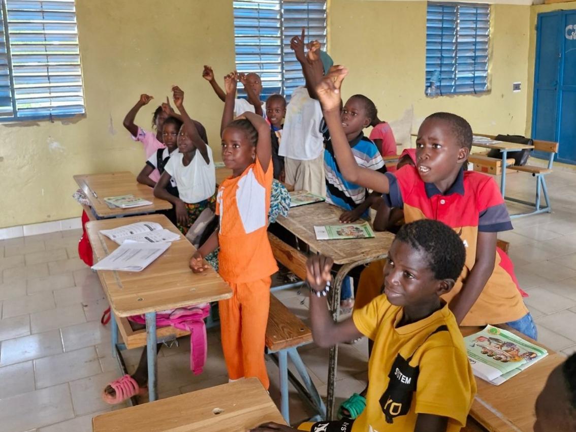 A group of children raising their hands in a classroomDescription automatically generated
